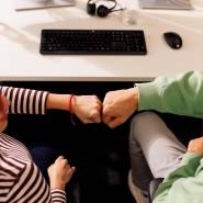 Kollege und Kollegin sitzen gemeinsam im Kaufland-Büro am Schreibtisch mit schwarzer Tastatur. Ihre zur Faust geballten Hände berühren sich zum Gruß.