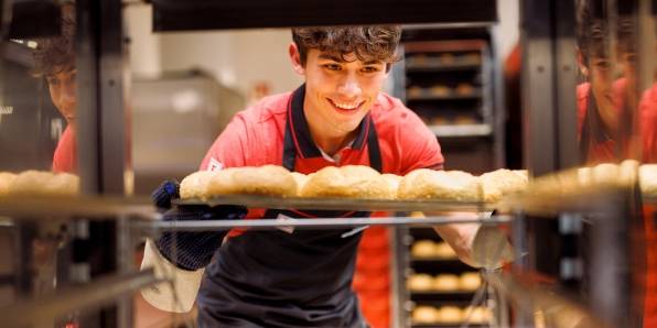 Junger Verkaufsmitarbeiter von Kaufland zieht ein Blech frischgebackener Brötchen aus dem Backregal.