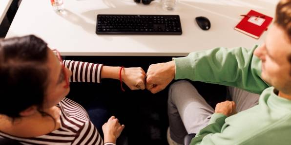 Kollege und Kollegin sitzen gemeinsam im Kaufland-Büro am Schreibtisch mit schwarzer Tastatur. Ihre zur Faust geballten Hände berühren sich zum Gruß.