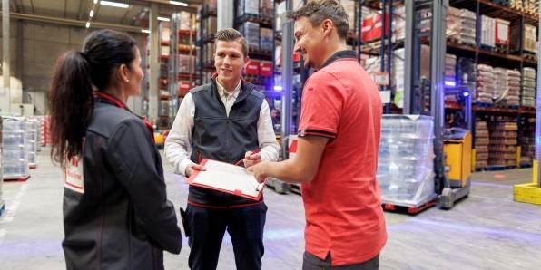 Logistik-Mitarbeiter von Kaufland unterhält sich im Lager mit seiner Kollegin und seinem Kollegen und macht sich Notizen auf seinem Clip-Board.