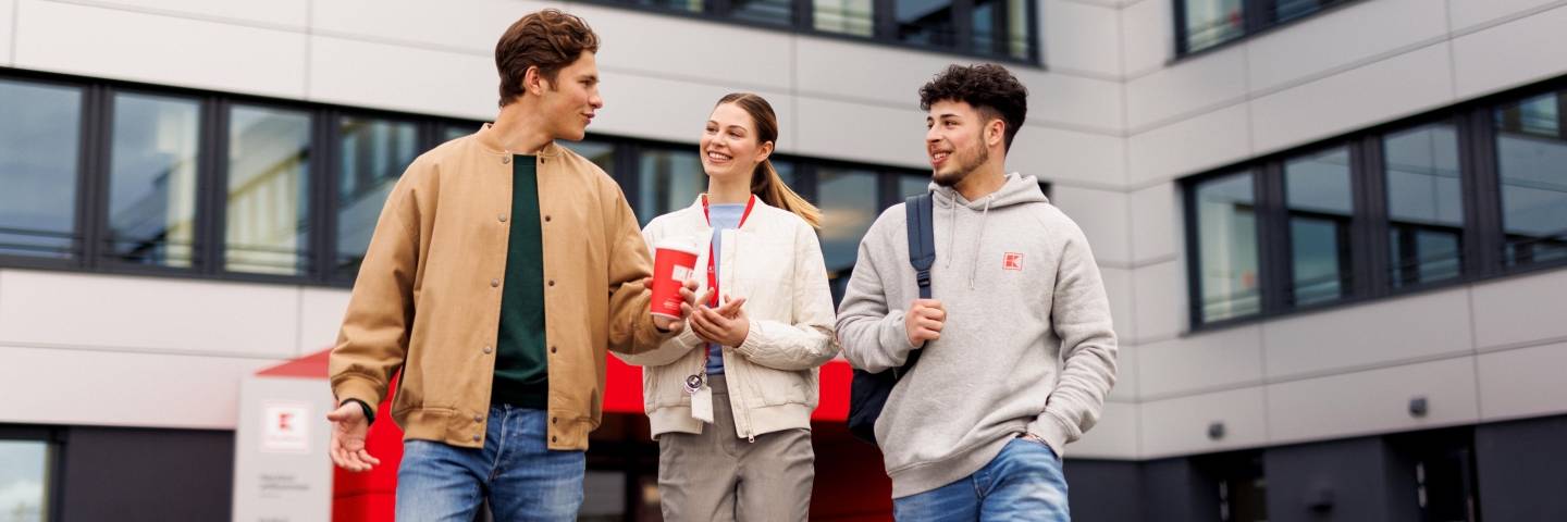 Drei Azubis stehen vor dem Haupteingang der Kaufland-Zentrale. Außen stehen zwei jungen Männer, in der Mitte eine junge Frau. Alle drei sind salopp gekleidet.