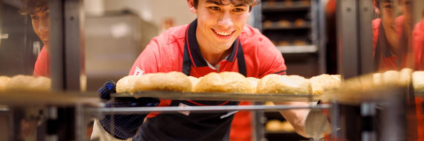 Junger Verkaufsmitarbeiter von Kaufland zieht ein Blech frischgebackener Brötchen aus dem Backregal.