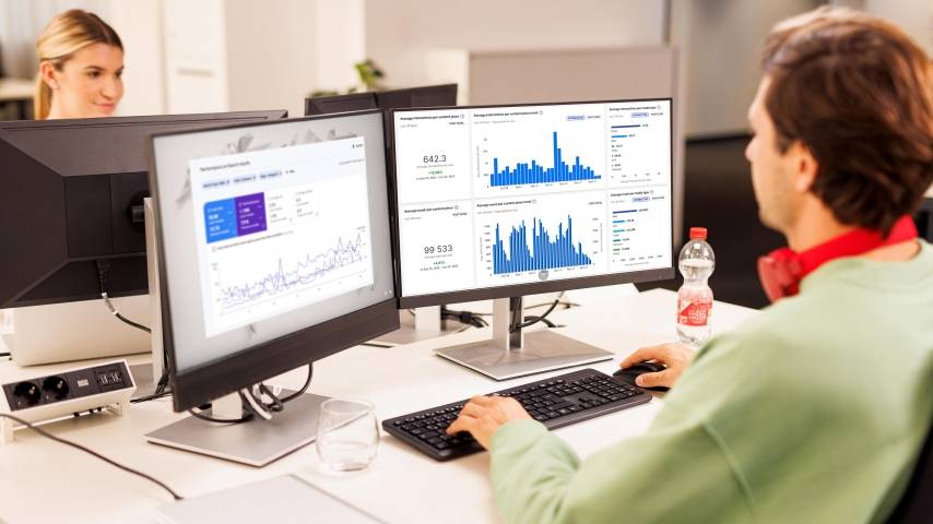 Kaufland-Mitarbeiter am Arbeitsplatz. Auf seinen beiden Bildschirmen sieht man Diagramme. Gegenüber von ihm sitzt eine Kollegin ebenfalls am Schreibtisch.
