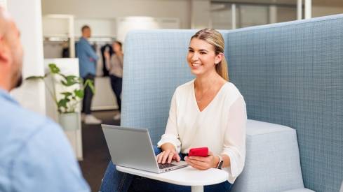 Kaufand-Mitarbeiterin in stylischer Büro-Lounge mit Laptop. Sie ist im Gespräch mit einem Kollegen.