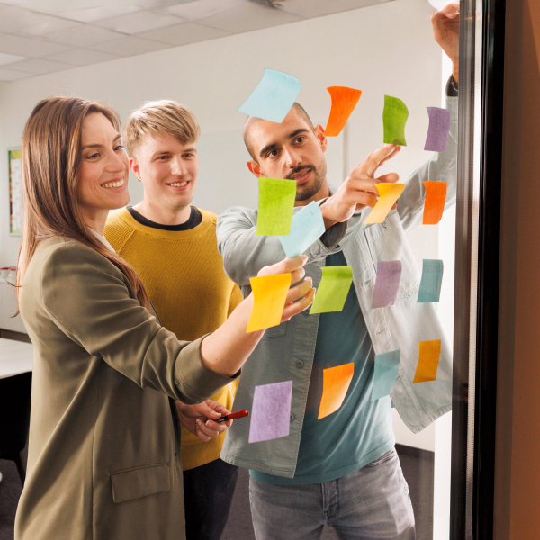 Eine junge Frau, ein junger Mann, beide Trainees und ein Ausbilder sind in einem Meeting. Die drei stehen vor einer gläsernen Wand und arbeiten mit Post-Its.