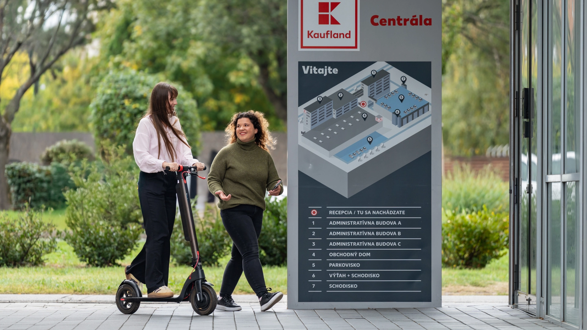 Študentka a brigádnička na kolobežke pred vchodom do centrály