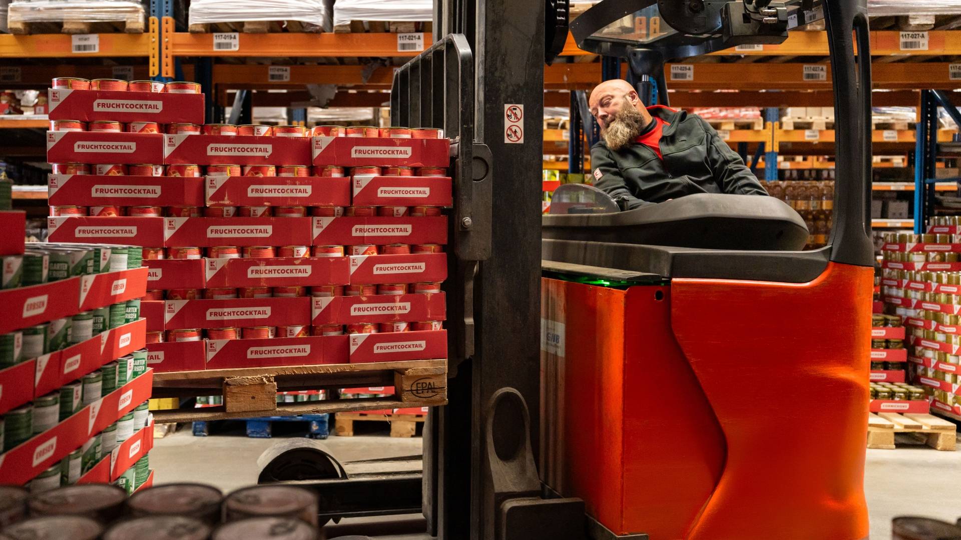 Skladník na červenom vysokozdvižnom vozíku naberá palety s konzervami