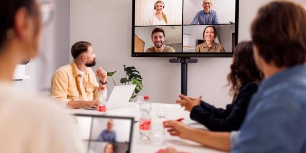 Hybride Zusammenarbeit: 4 Mitarbeiter im Besprechungsraum bei einem Termin mit anderen Kollegen, die über Teams auf einem großen Bildschirm zugeschaltet sind.